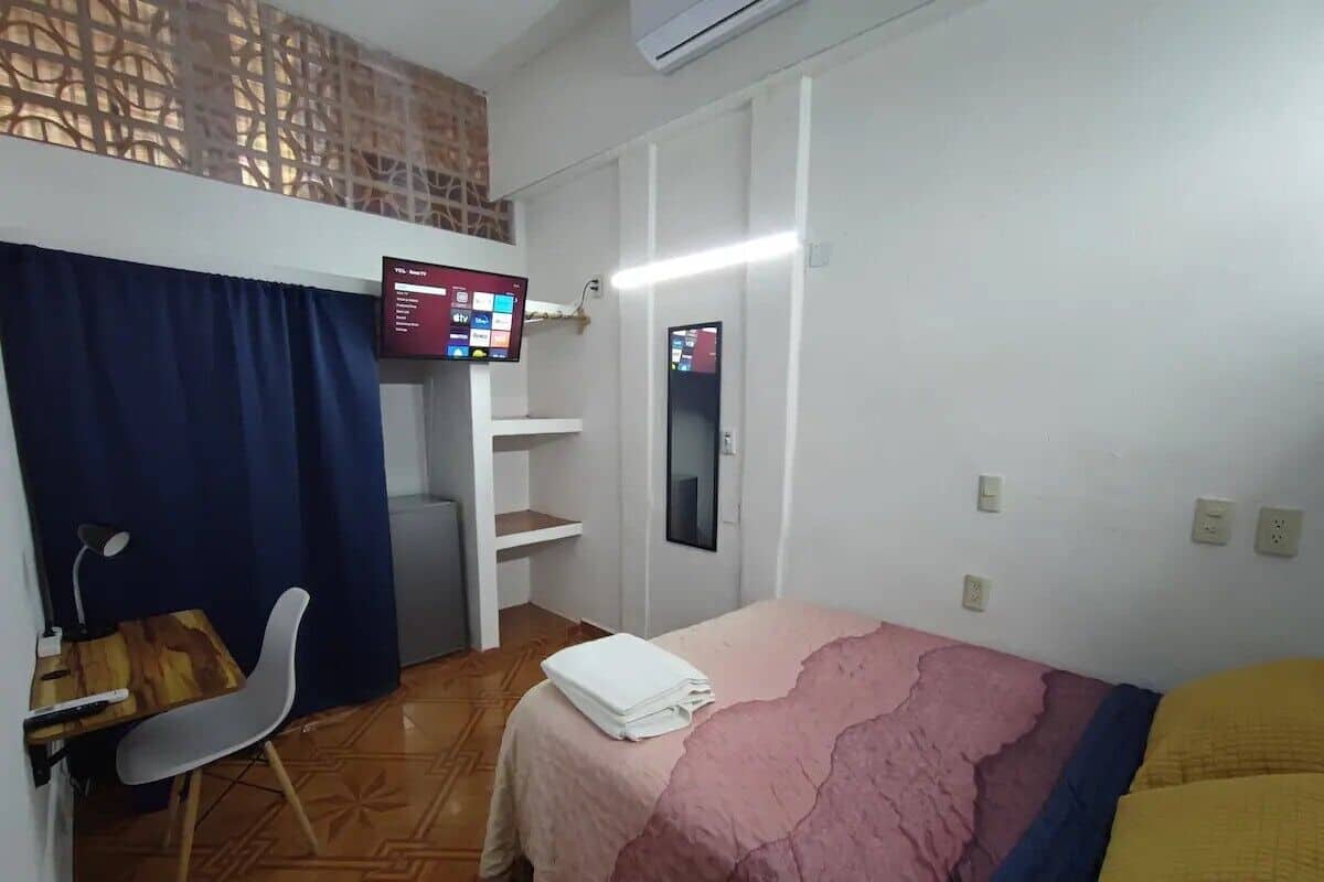 A room with a bed, a desk with a chair, a TV, and a shelf.
