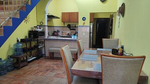 Μια κουζίνα με πλακάκια στο δάπεδο, ξύλινα ντουλάπια, έναν νεροχύτη και μια κουζίνα. Υπάρχει επίσης ένα τραπέζι φαγητού με καρέκλες και ένας διανομέας νερού.