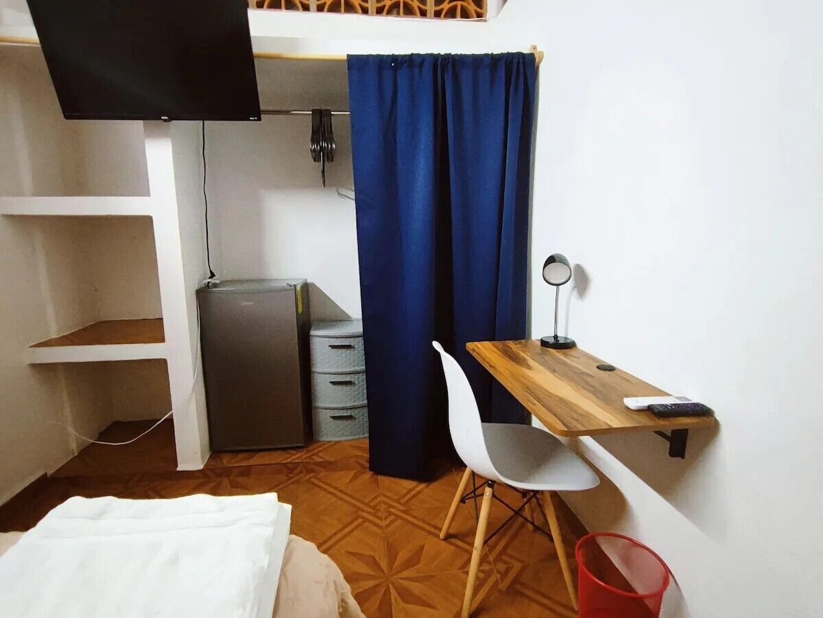 A compact room with a bed, a desk with a chair, a small refrigerator, a TV mounted on the wall, and a curtain.