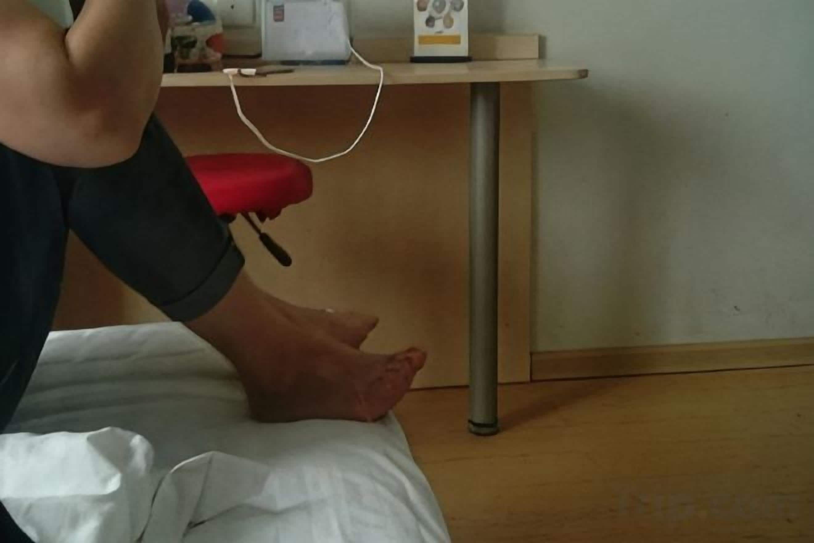 Una cama con sábana blanca, almohada roja y mesita de noche con un dispositivo blanco y una cajita.