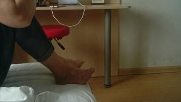 Una cama con sábana blanca, almohada roja y mesita de noche con un dispositivo blanco y una cajita.