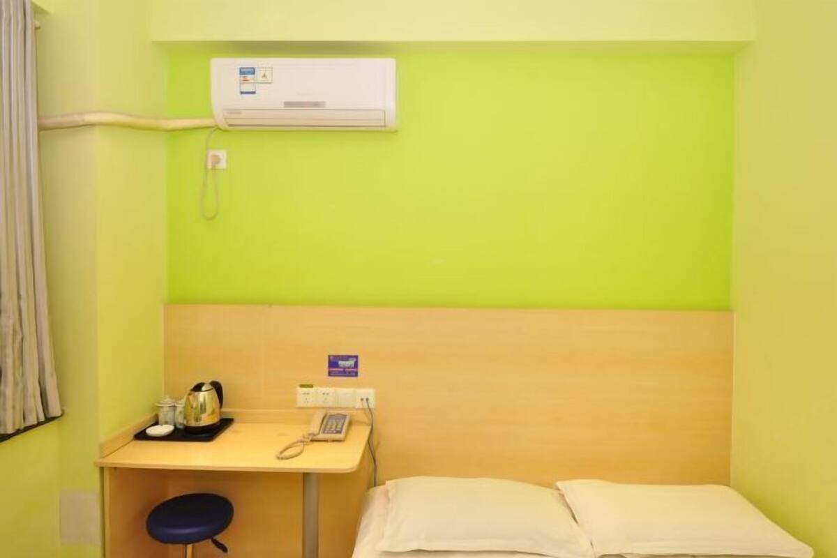 A small, single-room hotel room with a bed, a desk with a telephone, a kettle, and a cup.