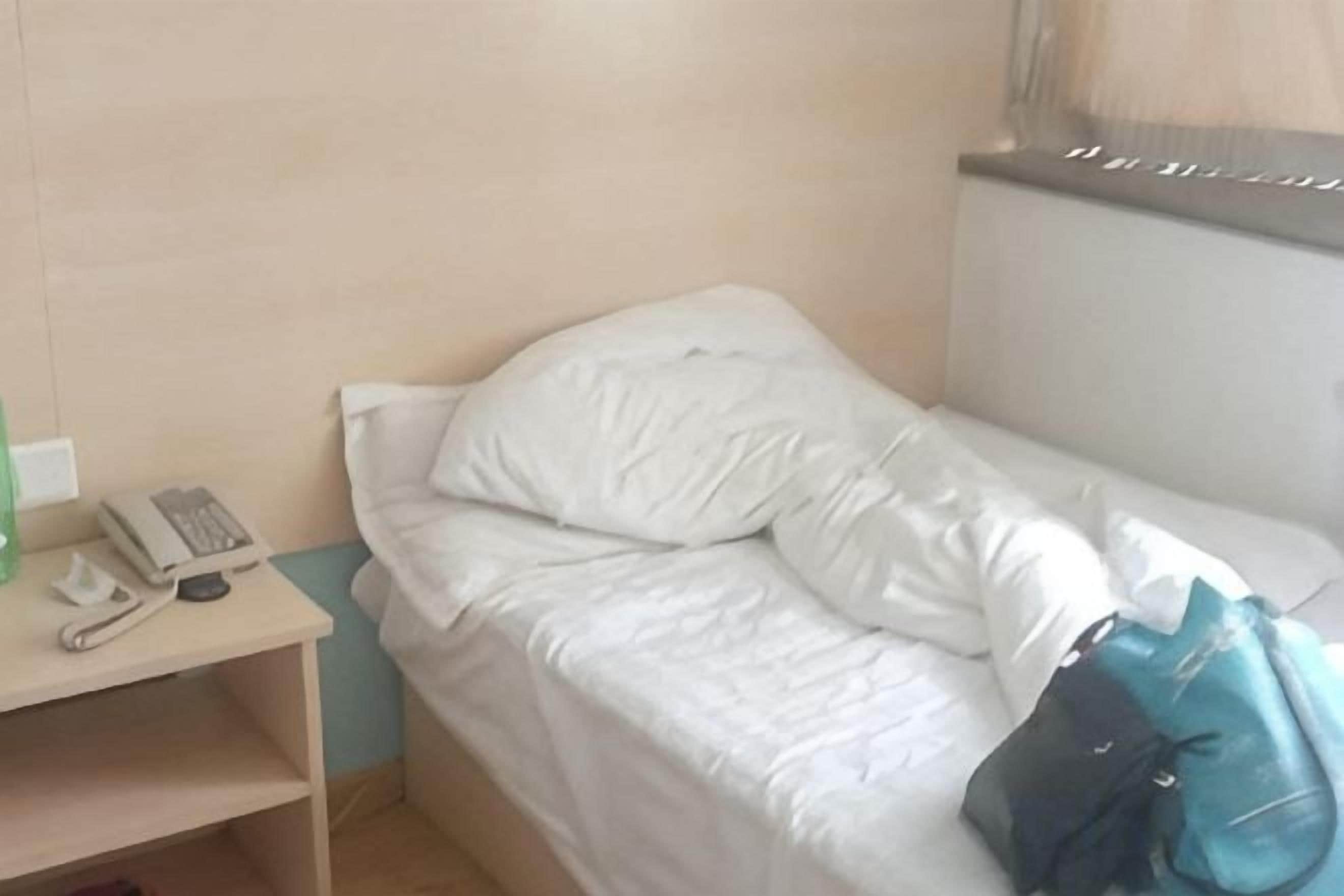 Un lit simple avec une literie blanche, une table de chevet avec un téléphone et un sac bleu posé sur le lit.