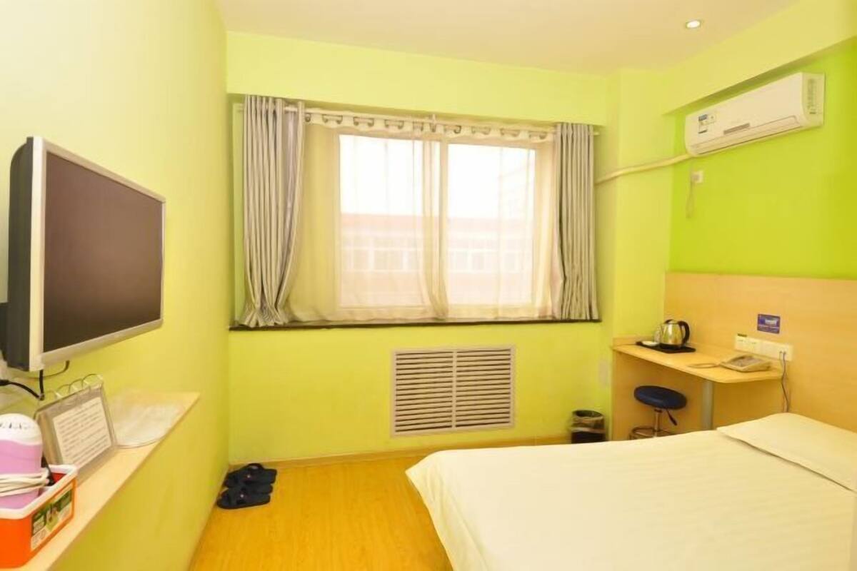 A hotel room with a bed, a desk with a kettle, a television, a window with curtains, and an air conditioning unit.
