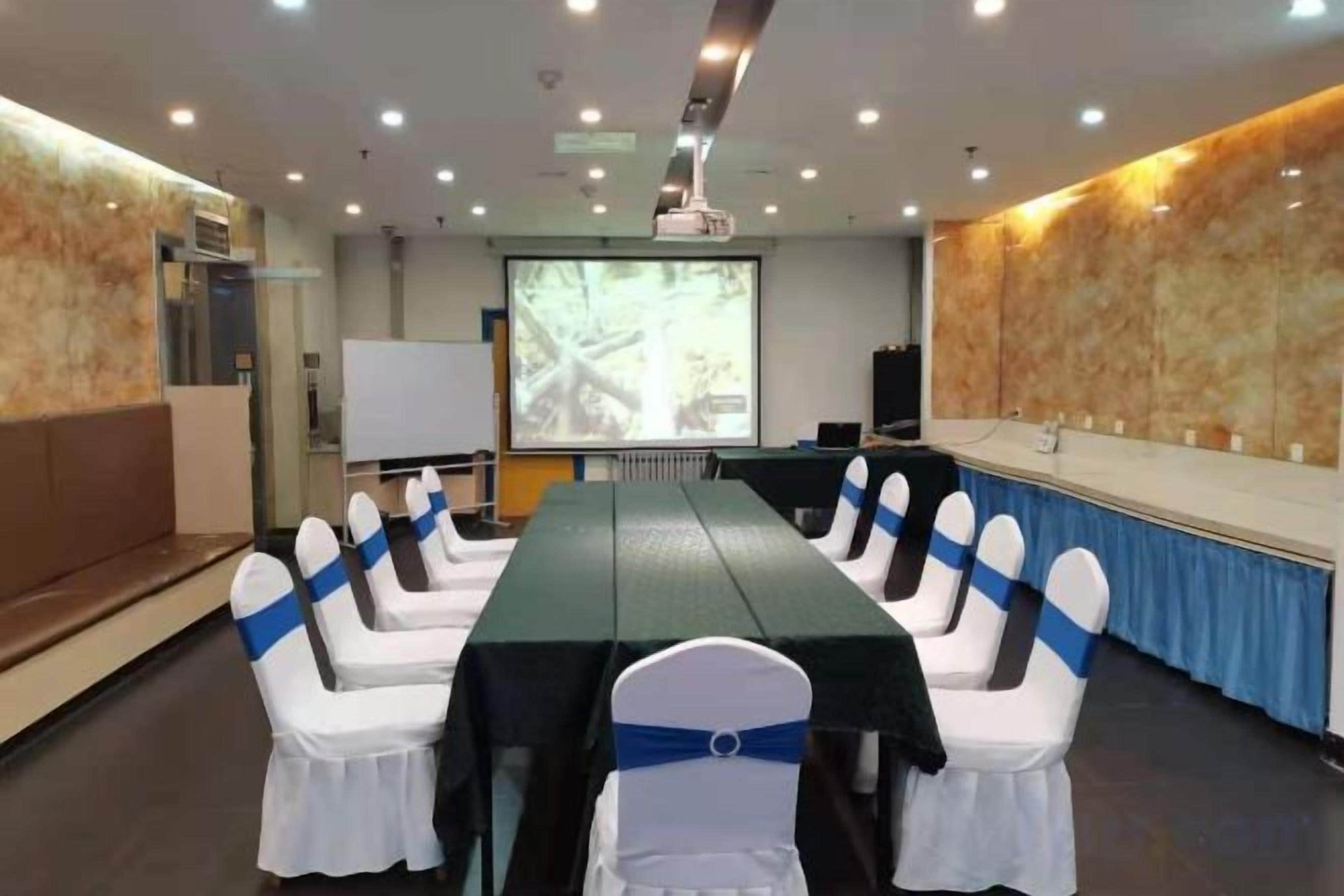 Une salle de conférence avec une longue table, des chaises blanches ornées de rubans bleus, un écran de projection et un tableau blanc.
