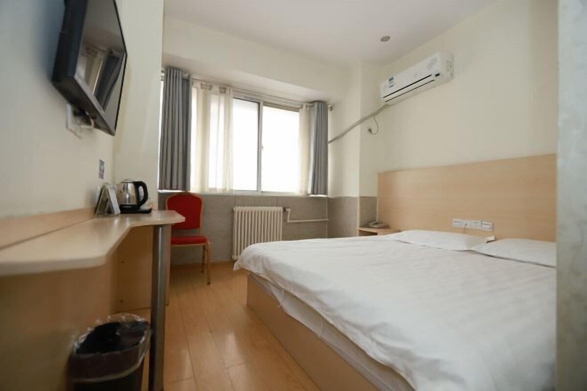 A hotel room with a bed, a desk, a chair, a TV, and an air conditioning unit.
