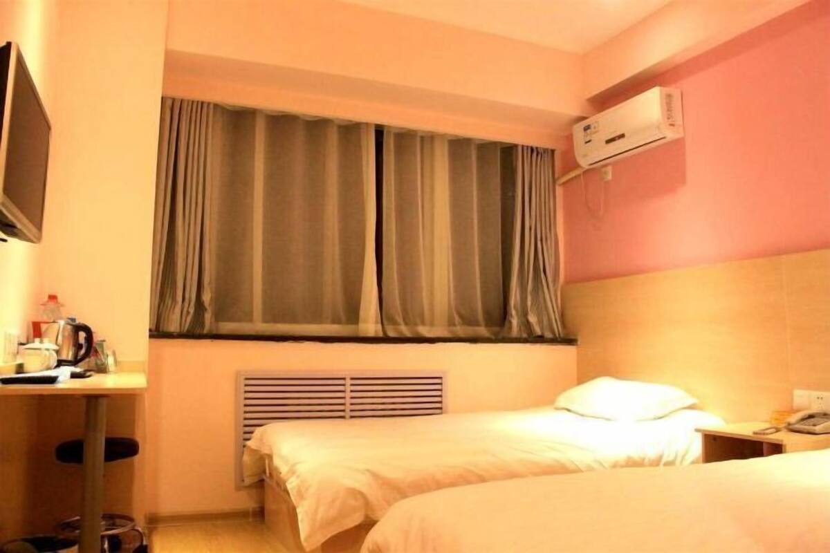 A hotel room with two beds, a TV, an air conditioner, a window with curtains, and a radiator.