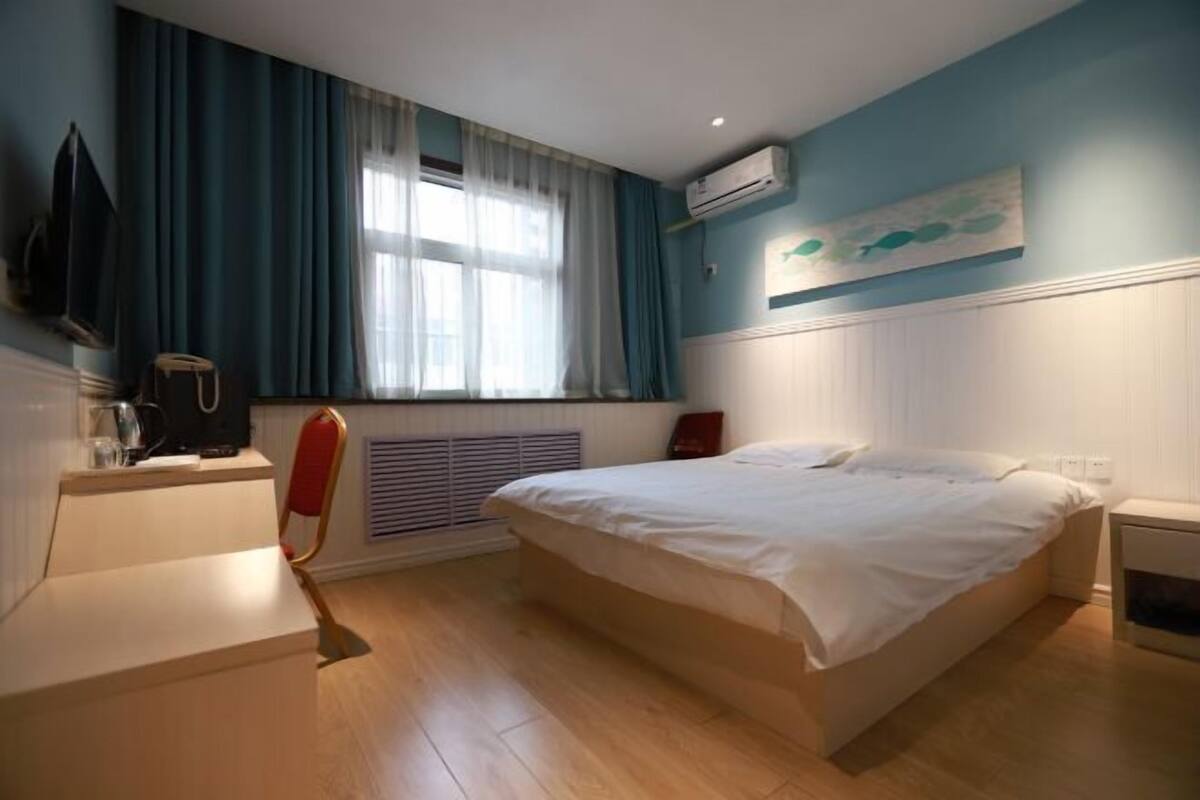 A bedroom with a bed, a chair, a heater, a window with curtains, an air conditioner, and a painting on the wall.