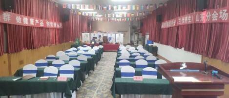 Un salón de conferencias con filas de mesas y sillas, un atril y cortinas rojas.
