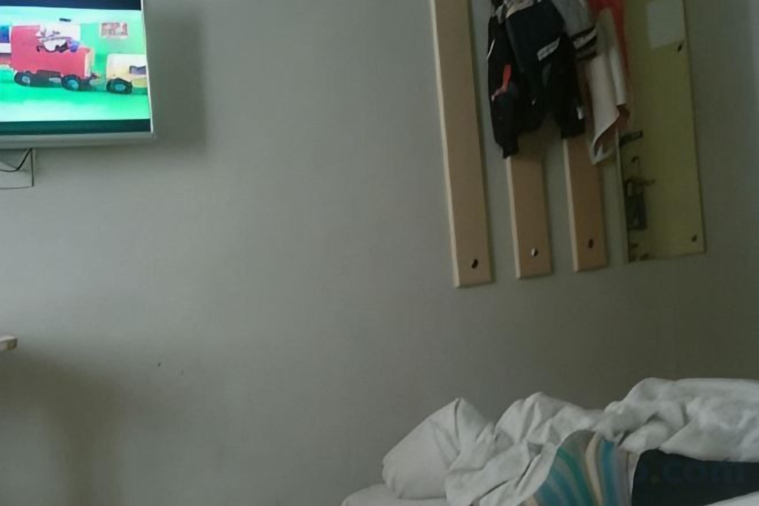 A hotel room with a bed, a television mounted on the wall, and a coat rack with hanging items.