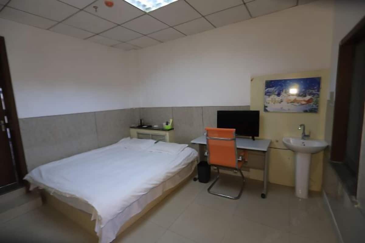 A bedroom with a bed, a desk, a chair, a sink, and a painting on the wall.