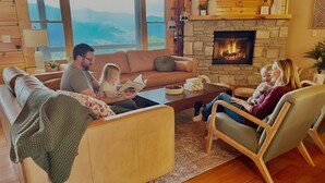 A cozy living room with a stone fireplace, large windows, and a view of mountains. - Hot tub, theater room w/100" tv, views, firepit, 3 king beds, porch perfection! (Maggie Valley)