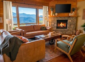 A cozy living room with a stone fireplace, leather sofas, and a large window offering a mountain view. - Hot tub, theater room w/100" tv, views, firepit, 3 king beds, porch perfection! (Maggie Valley)