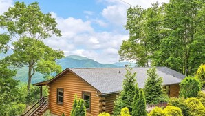 A log cabin with a steep staircase leading to the entrance, surrounded by lush greenery and mountains in the background. - Hot tub, theater room w/100" tv, views, firepit, 3 king beds, porch perfection! (Maggie Valley)