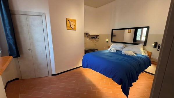 A hotel room with a bed, a door, a small table, a wall-mounted light, and a mirror.