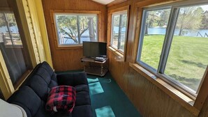 A cozy cabin interior with a dark-colored sofa, a television on a wooden stand, and large windows offering views of a grassy area and trees. - 2BR 1BA Cabin #8 at Riverside Point Resort (Park Rapids)