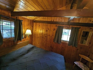 A wooden cabin room with a bed, a window with curtains, a lamp, and a small table.