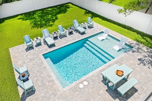 Piscina retangular com deck revestido, rodeada por cadeiras de lounge azuis claras e uma mesa com uma tigela.