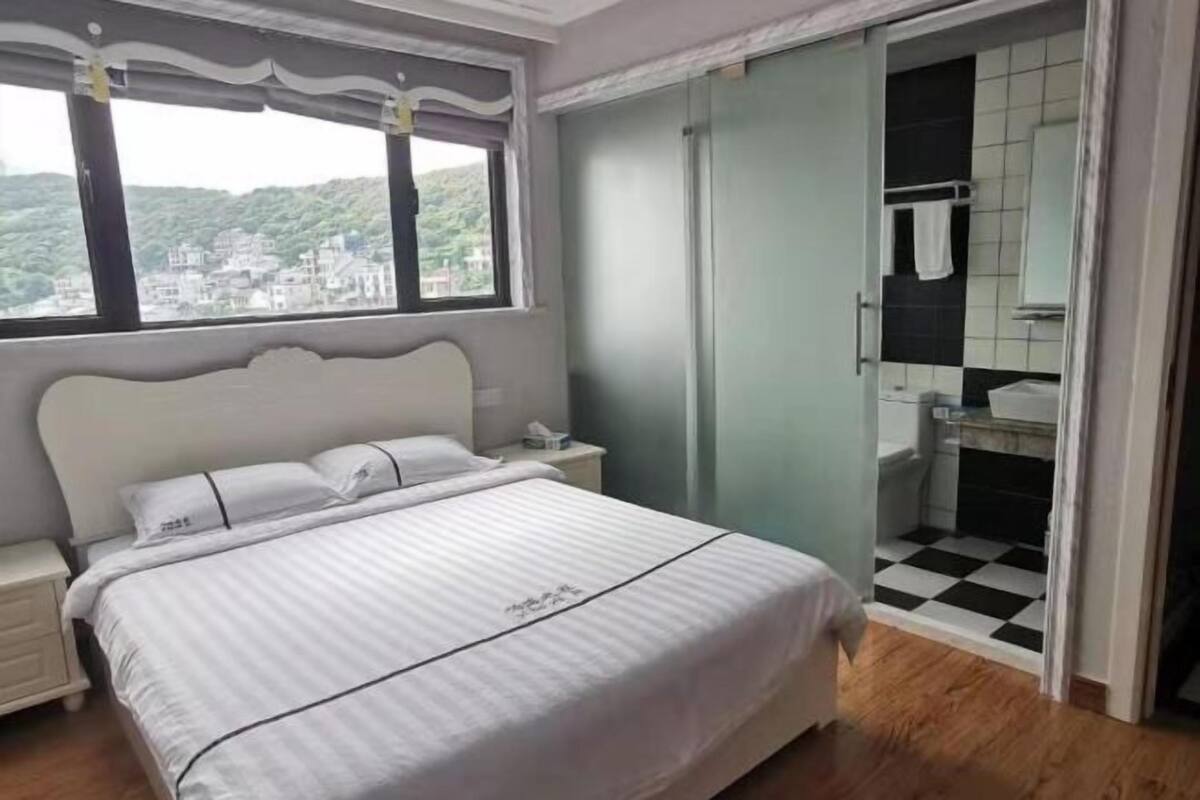 Ein Schlafzimmer mit Bett, Nachttisch, Fenster mit Aussicht, Badezimmer mit Waschbecken und Spiegel sowie einem karierten Boden.