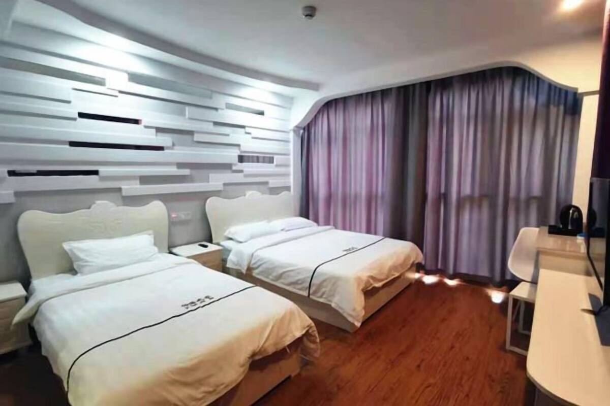 Ein Hotelzimmer mit zwei Betten, einem Schreibtisch, einem Stuhl, einem Fenster mit Vorhängen und einem modernen Wanddesign.