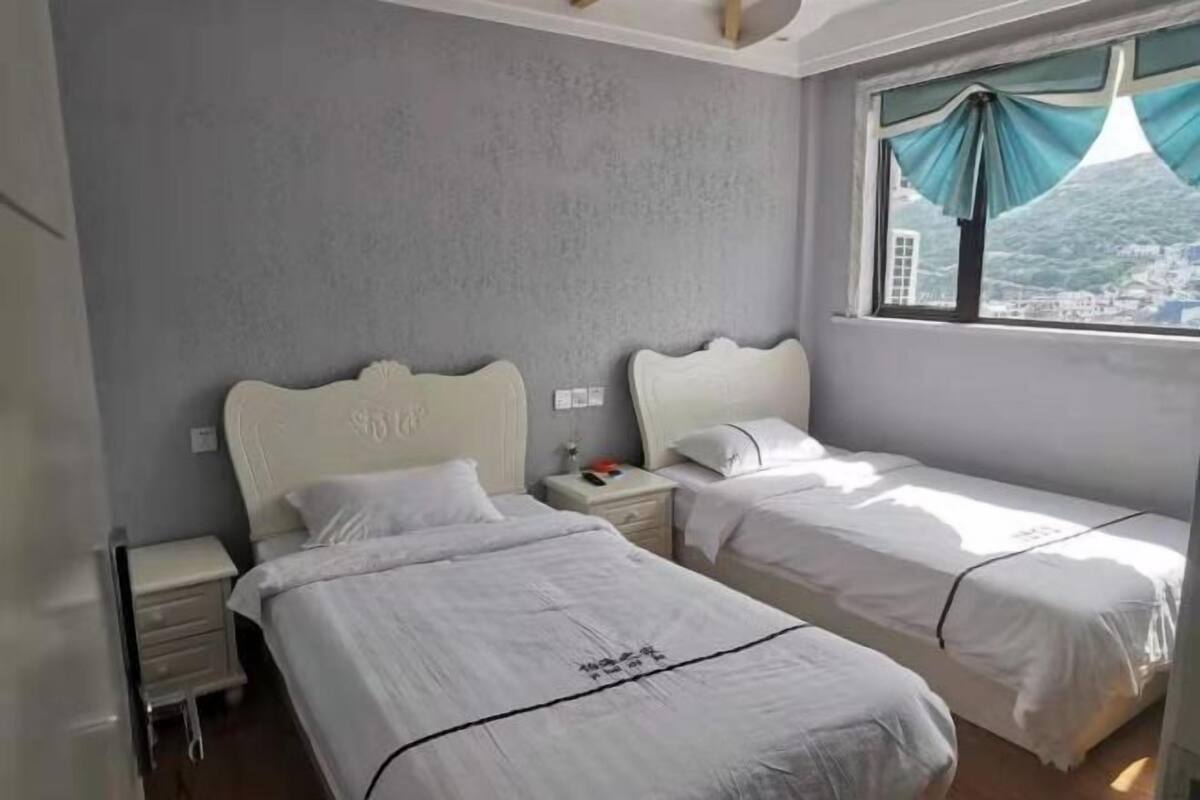 Ein Schlafzimmer mit zwei Betten, einem Fenster mit Vorhängen, einem Nachttisch und einer Wand mit strukturierter Oberfläche.