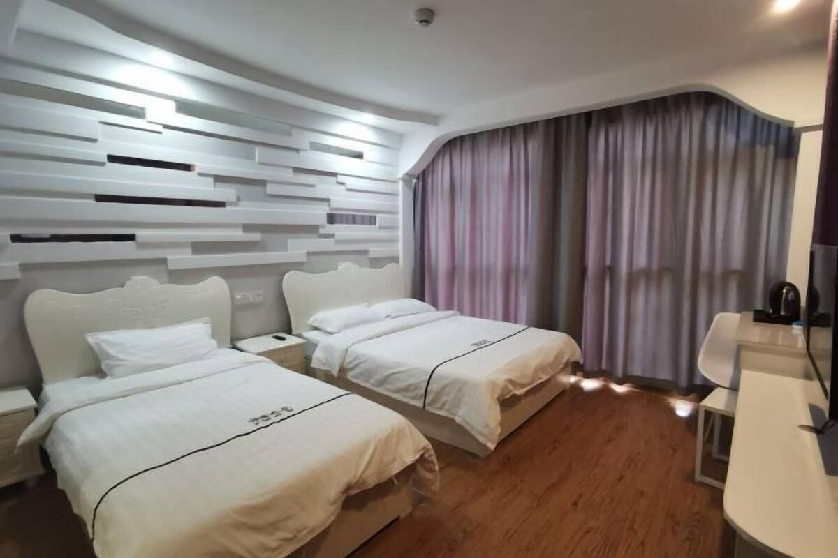 Ein Hotelzimmer mit zwei Betten, einem Schreibtisch, einem Stuhl, einem großen Fenster mit Vorhängen und einem modernen Wanddesign.