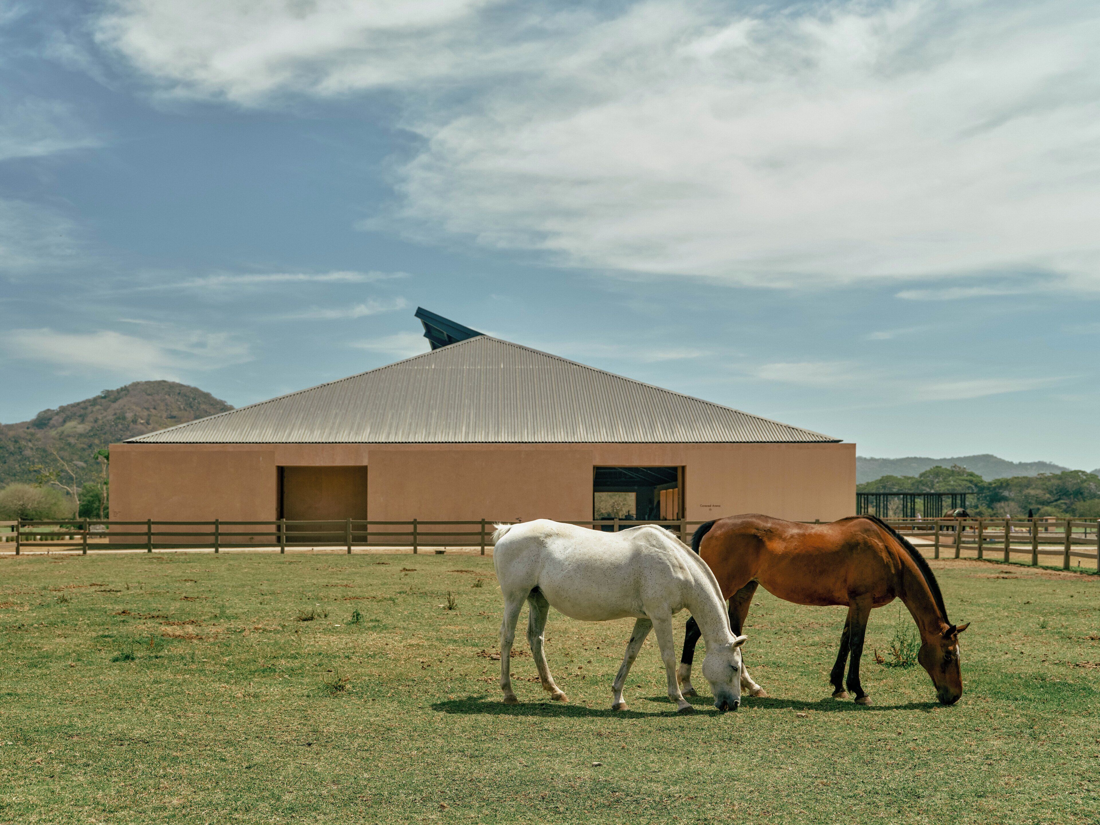 Un establo con dos caballos pastando en un campo cercado, con una montaña al fondo.