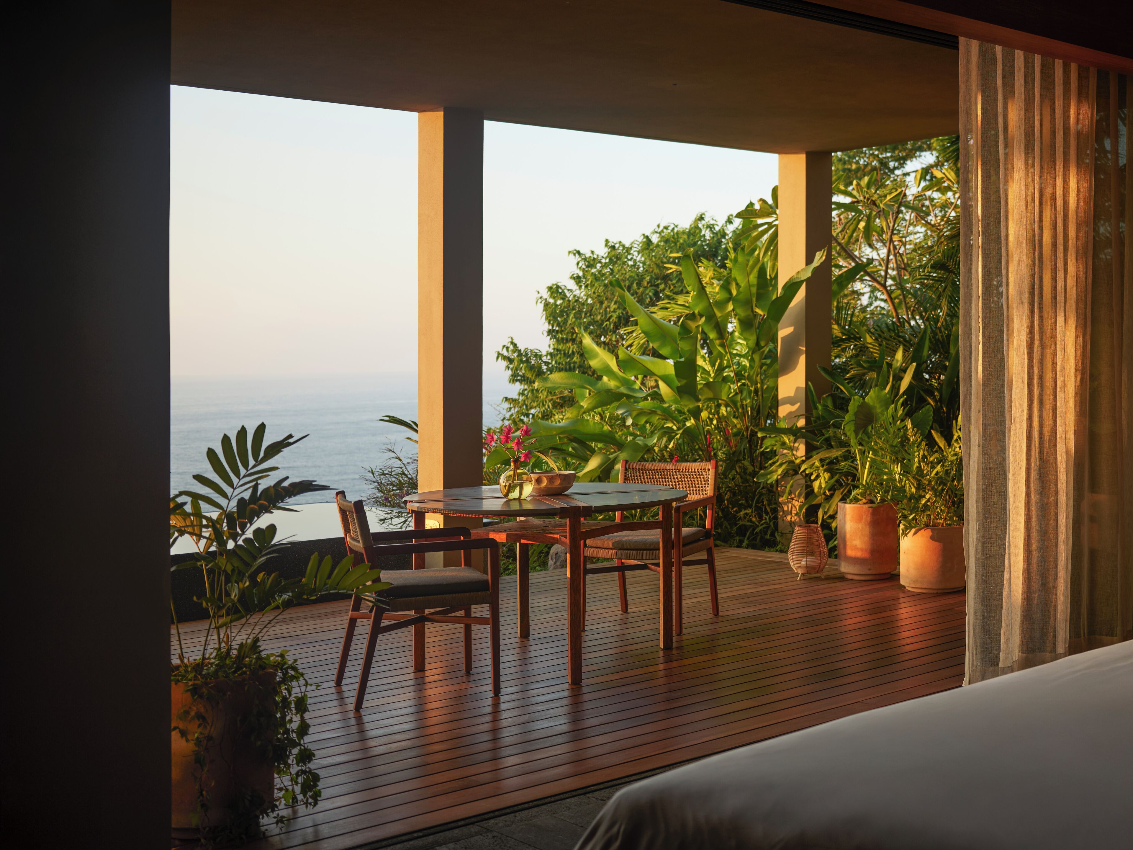 Varanda com mesa e cadeiras de madeira, plantas em vasos e vista para o mar.