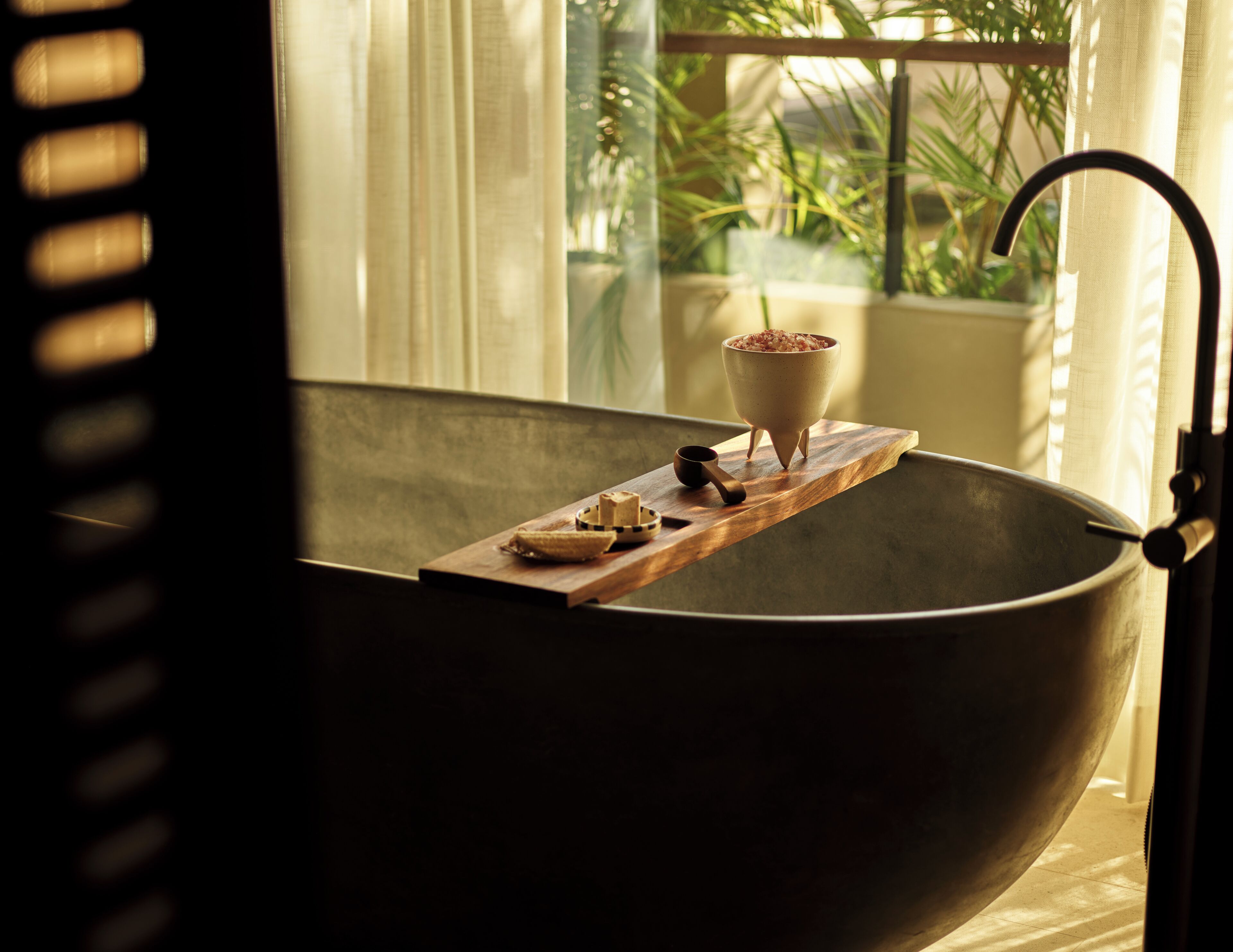 Una bañera grande de color oscuro con una bandeja de madera que sostiene un recipiente con sales de baño y una taza pequeña.