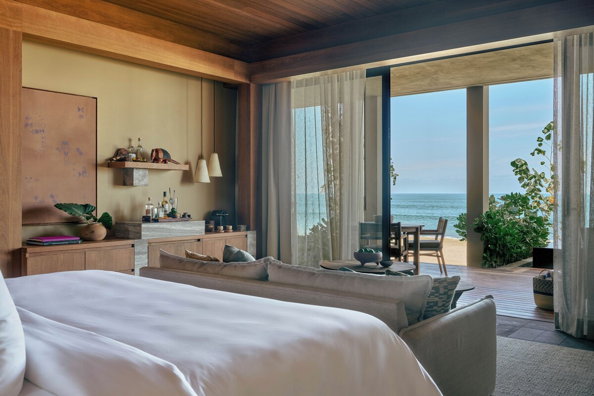 A modern hotel room with a large bed, a desk with a computer, a view of the sea, and a balcony with plants.