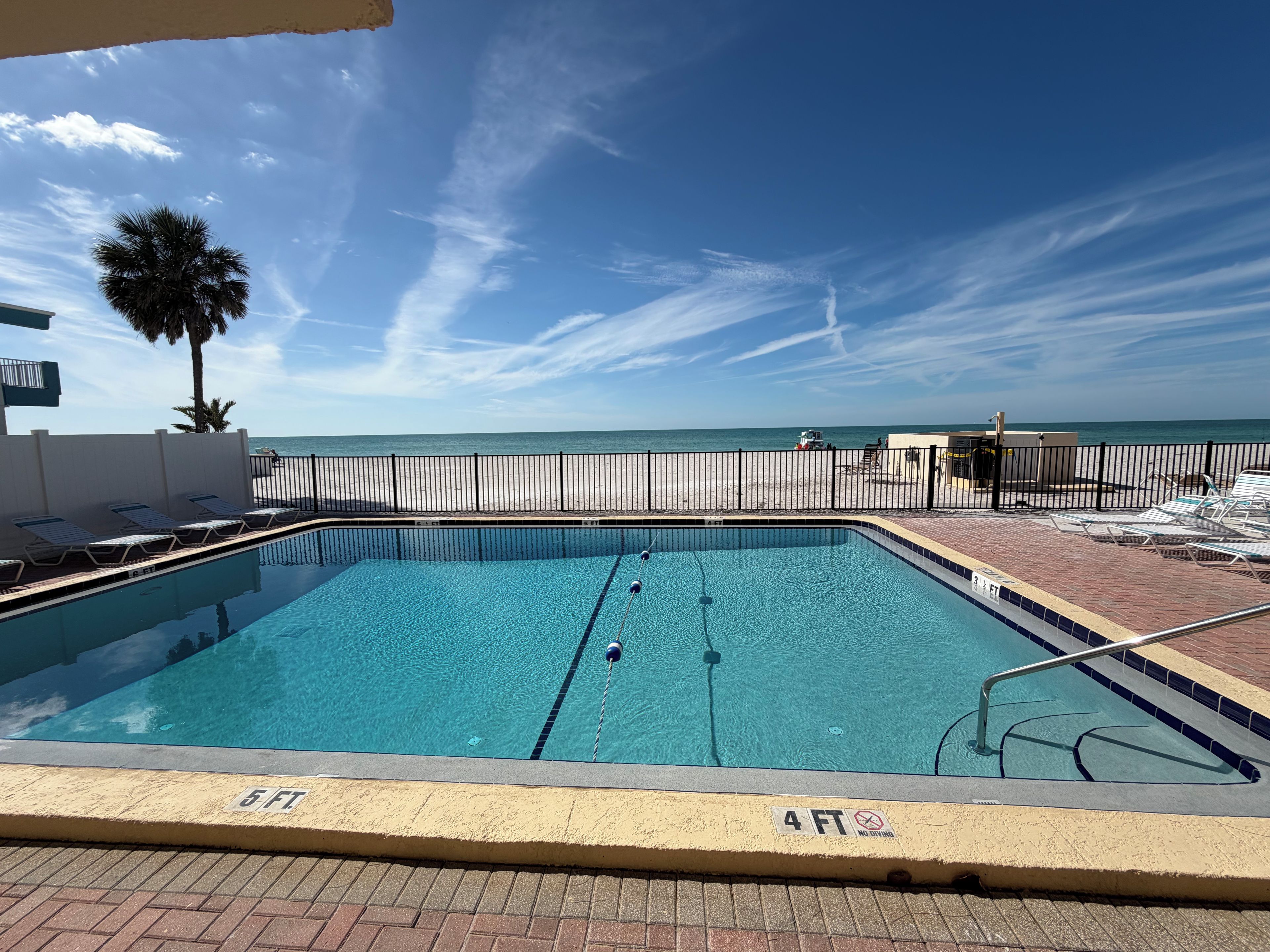 Ein rechteckiger Pool mit Sprungbrett und Rettungsschwimmerstand, umgeben von einem Zaun, mit Strand und Blick auf den Ozean im Hintergrund.