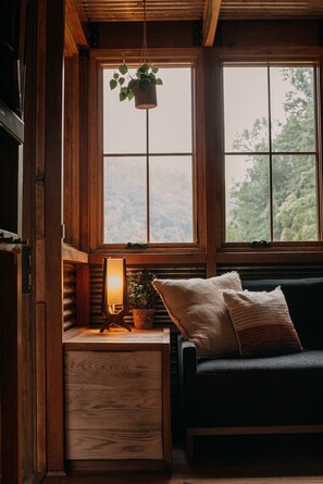 Una habitación acogedora con detalles de madera, una ventana con vistas a árboles, una planta colgante, una lámpara y un sofá con cojines.