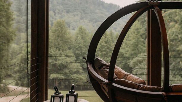 A wooden deck with a hanging chair and ottoman, overlooking a forested hillside.
