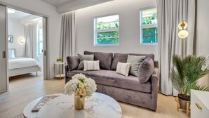 A modern living room with a grey sofa, a round coffee table with a vase of flowers, and a view of a garden through a window. - Vonder Miami Beach - Unit 3 (Miami Beach)