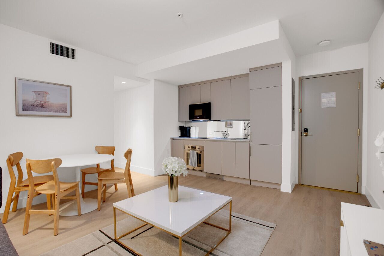 A modern kitchen with a microwave, oven, and sink. There is a dining area with a white table and wooden chairs. A framed picture hangs on the wall.