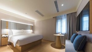 Una habitación de hotel moderna con una cama grande, una mesa de centro redonda, un sofá con cojines, un televisor y una ventana con cortinas.