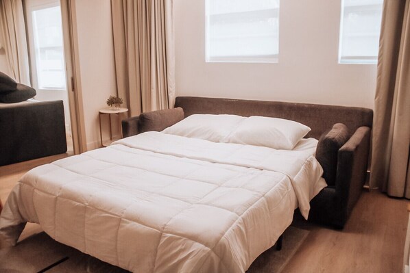 A neatly made bed with a white duvet, a dark headboard, and a small bedside table with a plant.
