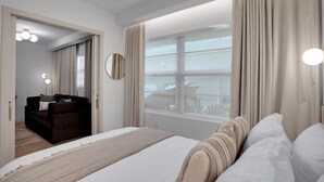Une chambre d’hôtel moderne dotée d’un grand lit, d’un coin salon avec un canapé et un fauteuil, d’un miroir et offrant une vue sur la ville par la fenêtre.