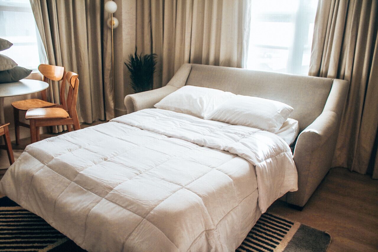 A neatly made bed with a white duvet and pillows, a wooden chair, a small round table, and a potted plant.