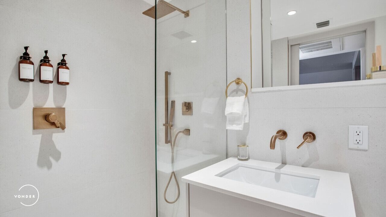 Moderni kylpyhuone, jossa on lasinen suihkuseinä, valkoinen pesuallas ja kolme shampoopulloa seinällä.