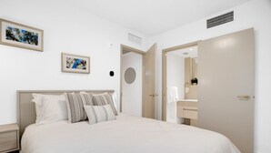 Une chambre à coucher comprenant un lit, une table de chevet, une bouche d’aération fixée au mur et une porte donnant accès à une salle de bain.