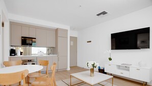 A modern kitchen with white cabinets, a built-in oven, and a microwave. There is a dining area with a white table and wooden chairs. A flat-screen TV is mounted on the wall above a white console table. - Vonder Miami Beach - Unit 14 (Miami Beach)