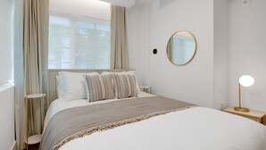 A bedroom with a large bed, a bedside table with a lamp, a round mirror on the wall, and a window with curtains. - Vonder Miami Beach - Unit 16 (Miami Beach)