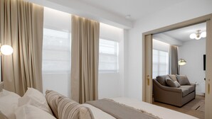 Moderni hotellihuone, jossa on suuri sänky, beigenväriset verhot, sohva ja näkymä kaupunkiin.