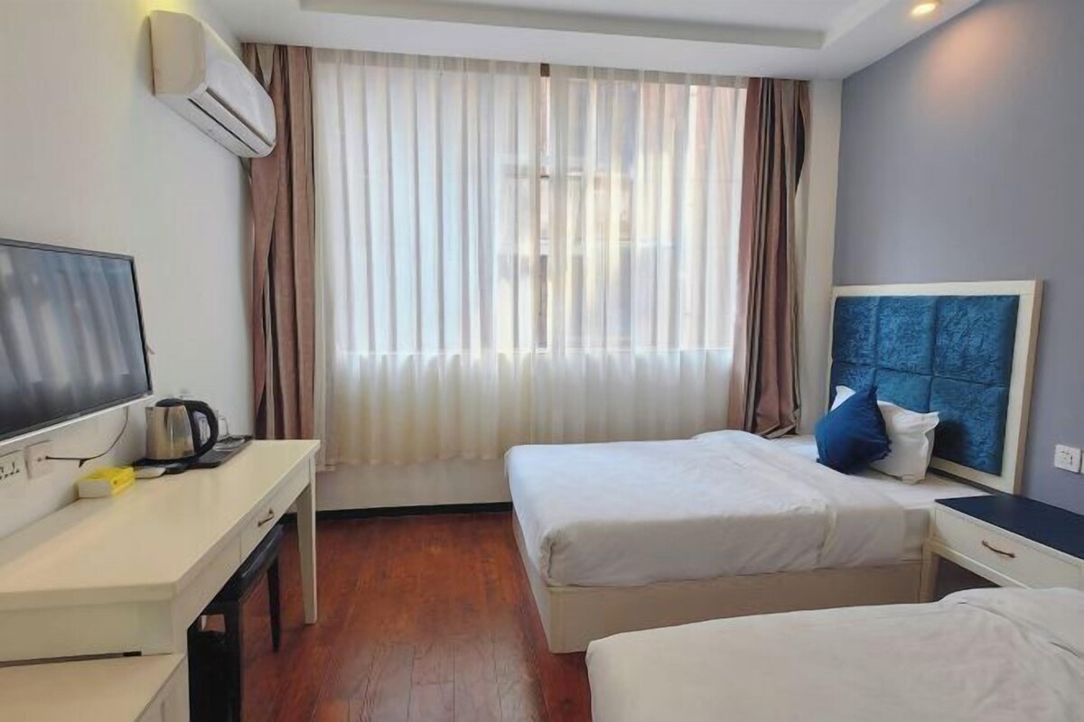 Quarto de hotel com duas camas, uma escrivaninha com televisão e cafeteira, e uma janela grande com cortinas.