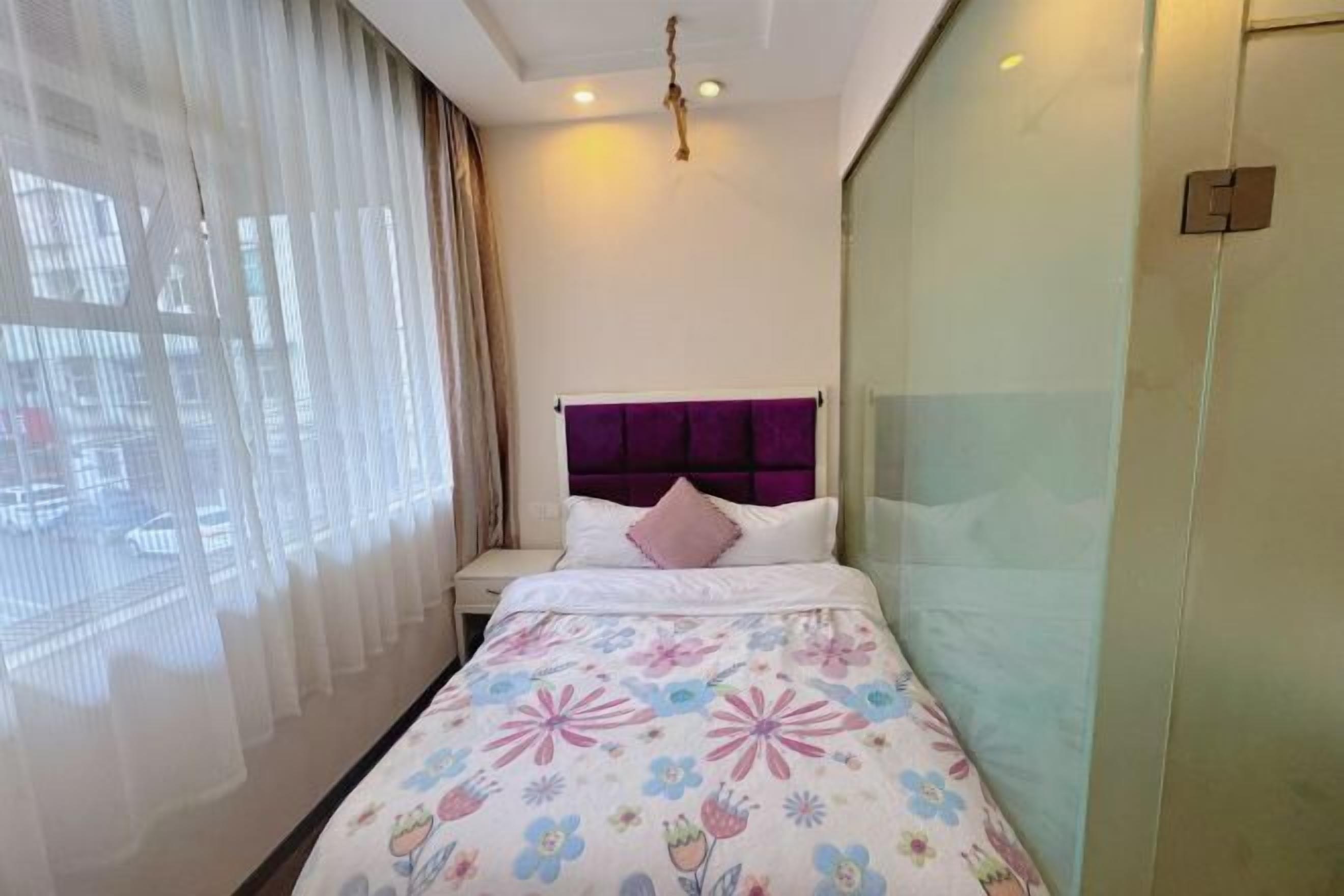 Habitación de hotel con cama, mesita de noche, ventana con cortinas translúcidas y lámpara de pared.