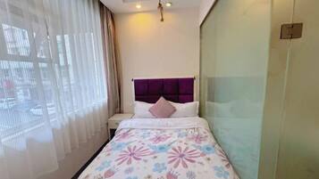 Habitación de hotel con cama, mesita de noche, ventana con cortinas translúcidas y lámpara de pared.