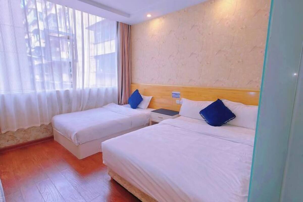 Una habitación de hotel con dos camas individuales, un cabecero de madera, una mesita de noche y una ventana con cortinas de gasa.