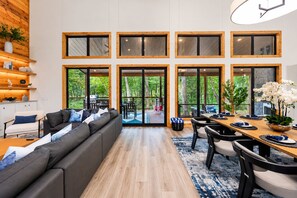 Un salon moderne avec un canapé, une table à manger et des chaises. De grandes fenêtres à cadre en bois offrent une vue sur la verdure environnante.