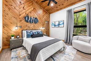 Une chambre avec un plafond en bois, un lit doté d’un matelas matelassé, un canapé gris, une table de chevet avec une horloge et une fenêtre avec des rideaux.